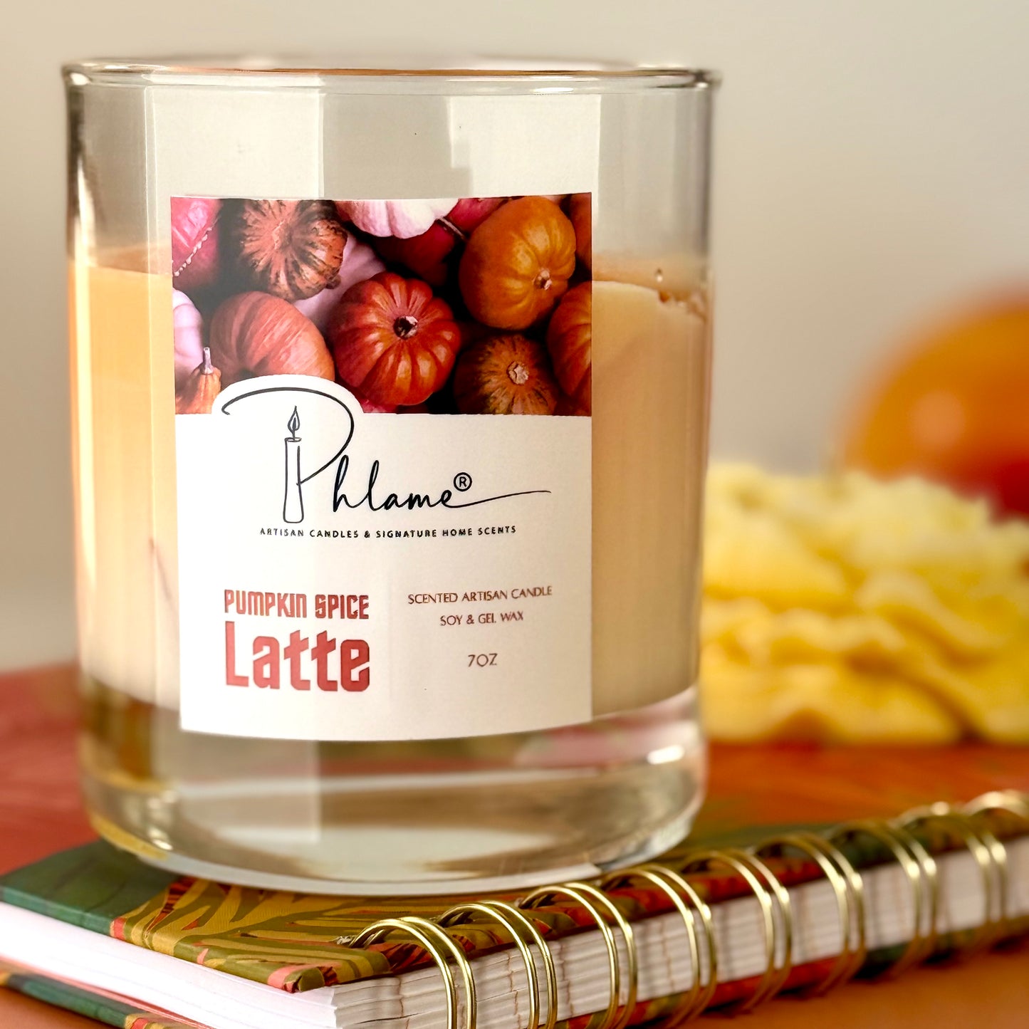 Candle labeled 'Pumpkin Spice Latte' by Phlame on a surface with pumpkins in the background.