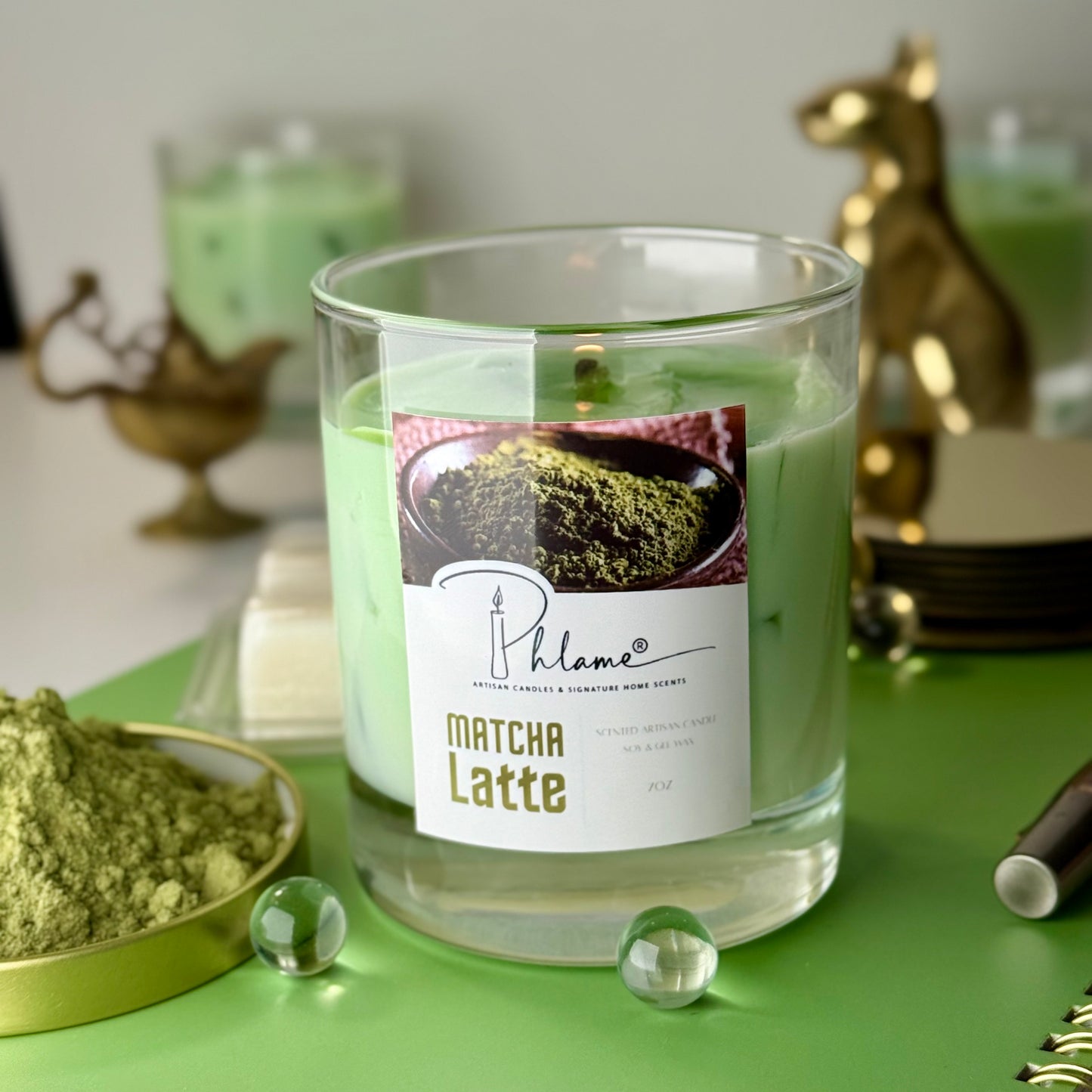 Matcha Latte candle with a label on a green surface, surrounded by matcha powder and decorative items.