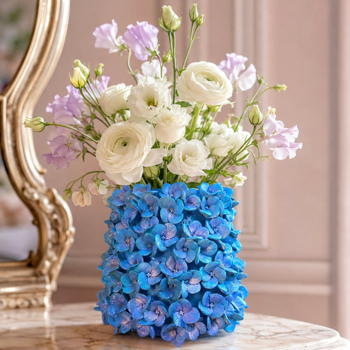 A vibrant blue hydrangea-covered vase filled with white ranunculus and light purple stock flowers, displayed on a marble side table next to an ornate gold mirror.