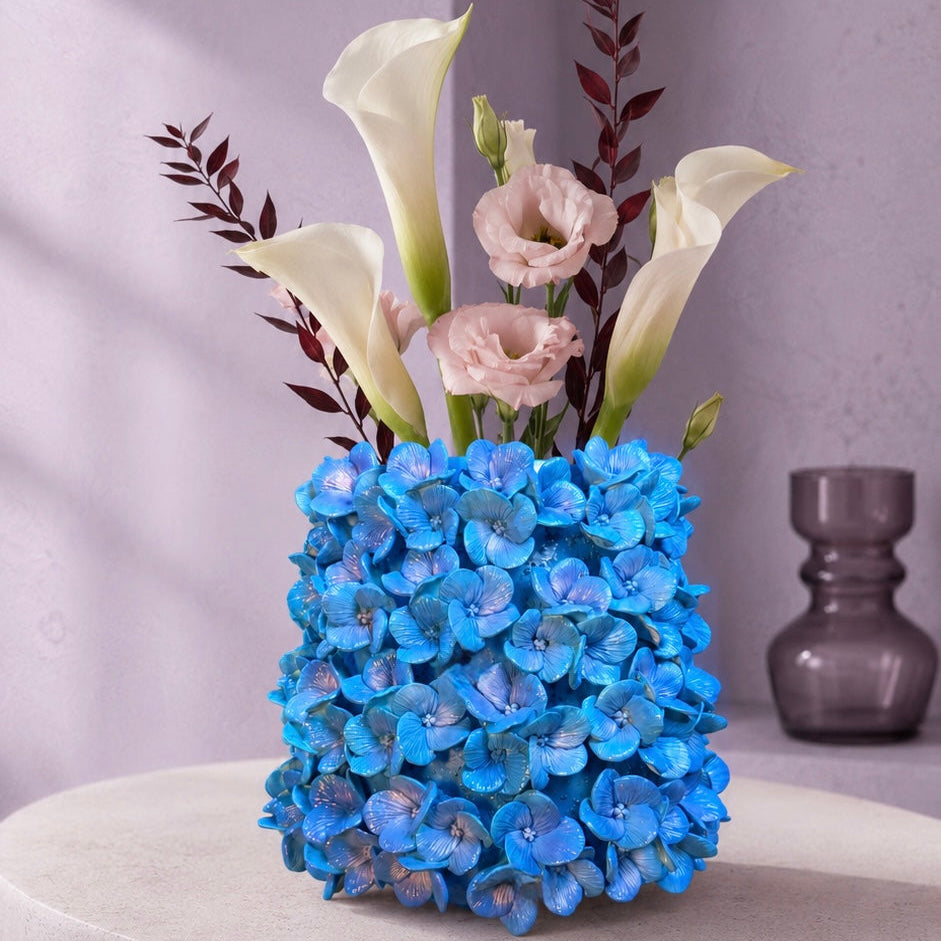 A floral arrangement featuring bright blue hydrangeas, white calla lilies, and soft pink roses in a textured cylindrical vase, displayed on a round pedestal against a lavender arched wall.