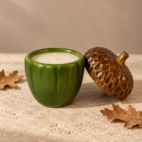A green ceramic candle vessel with a wick and white wax, shown beside a brown acorn-shaped lid on a textured beige surface with dried oak leaves.