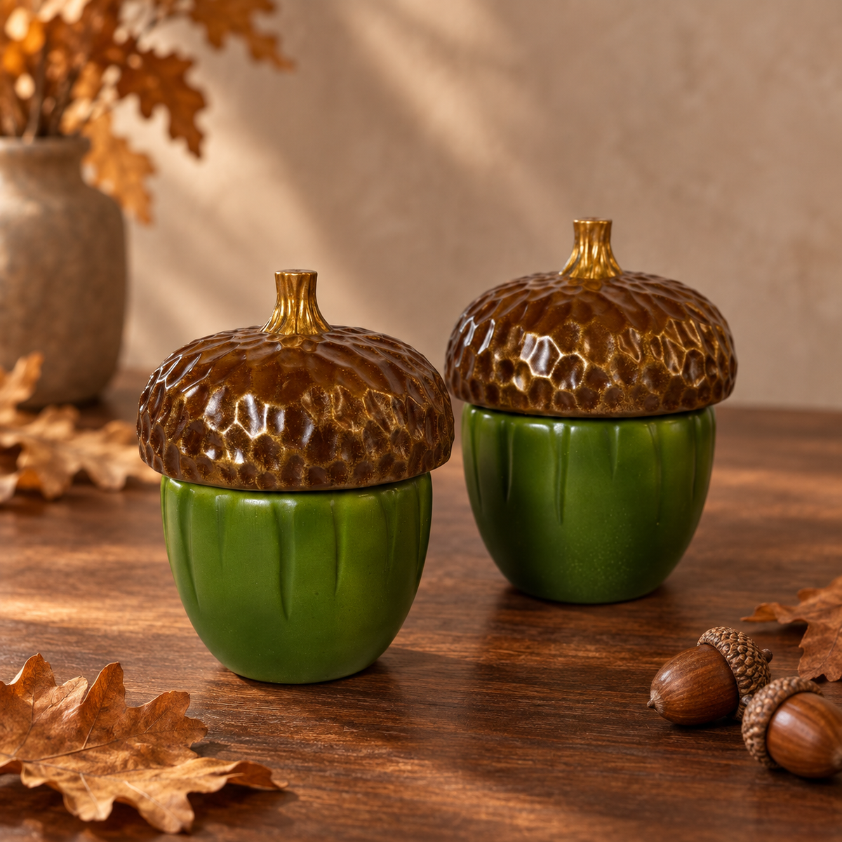 Two ceramic acorn-shaped containers with glossy green bodies and textured brown lids, displayed on a wooden surface with autumn leaves and a vase in the background.