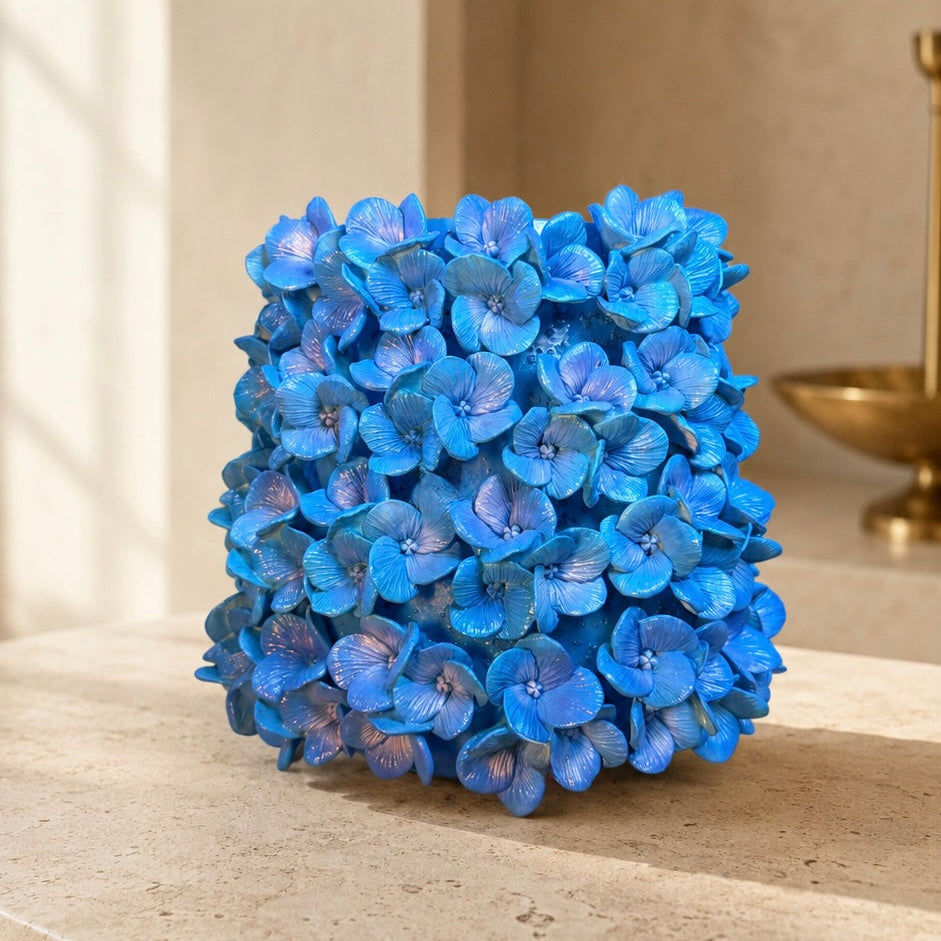 A decorative sculptural vase made of dense blue hydrangea blooms, displayed on a beige stone ledge in a warm, sunlit interior setting.