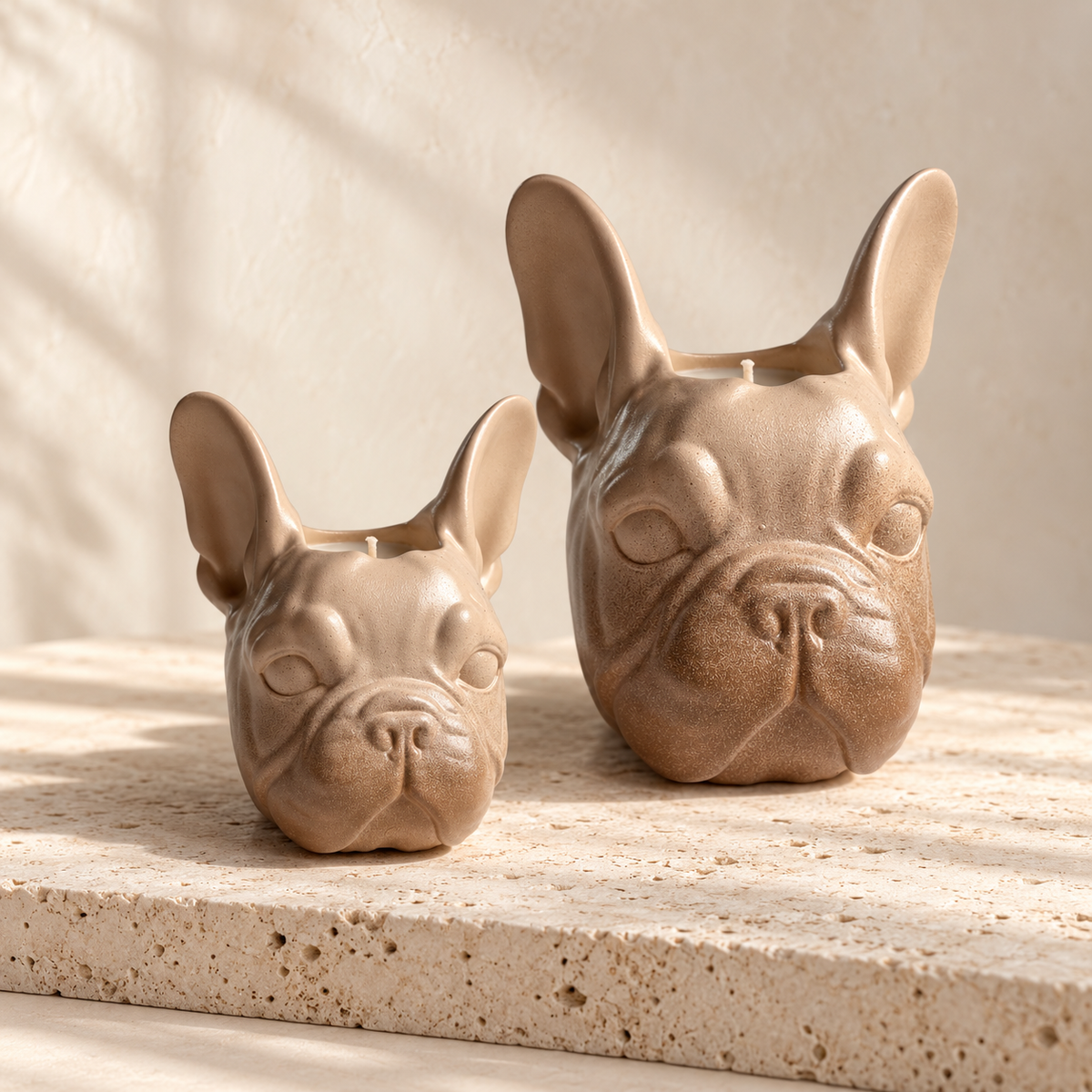 Two ceramic French bulldog-shaped candle holders in a warm beige and brown glaze, displayed on a textured stone surface with soft shadow overlays.