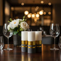 Decorative candles with gold stripes on a table with wine glasses and flowers in the background.