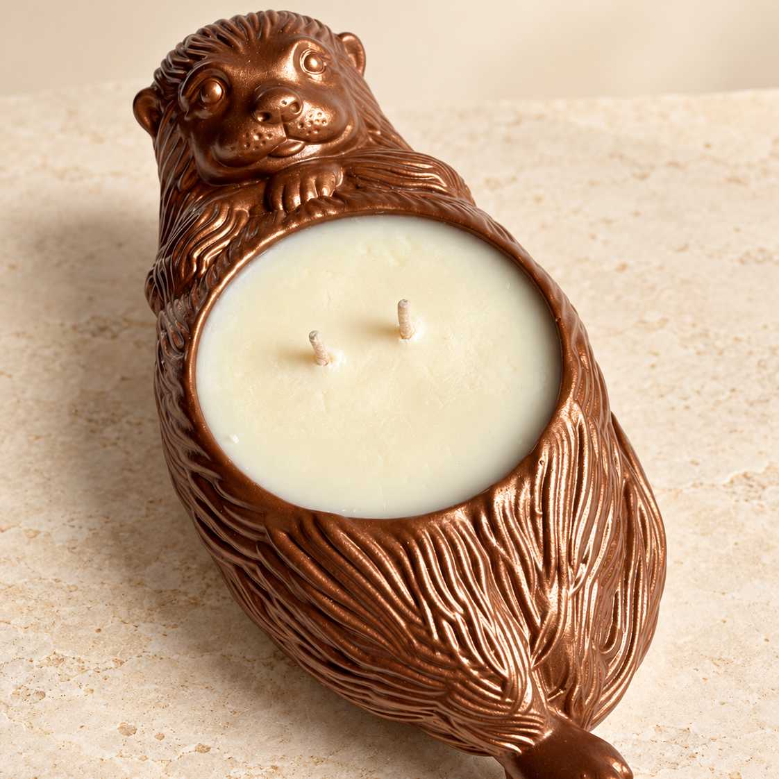 An otter-shaped candle holder shaped like a brown otter with a cream-colored wax candle inside, set against a light beige stone background.