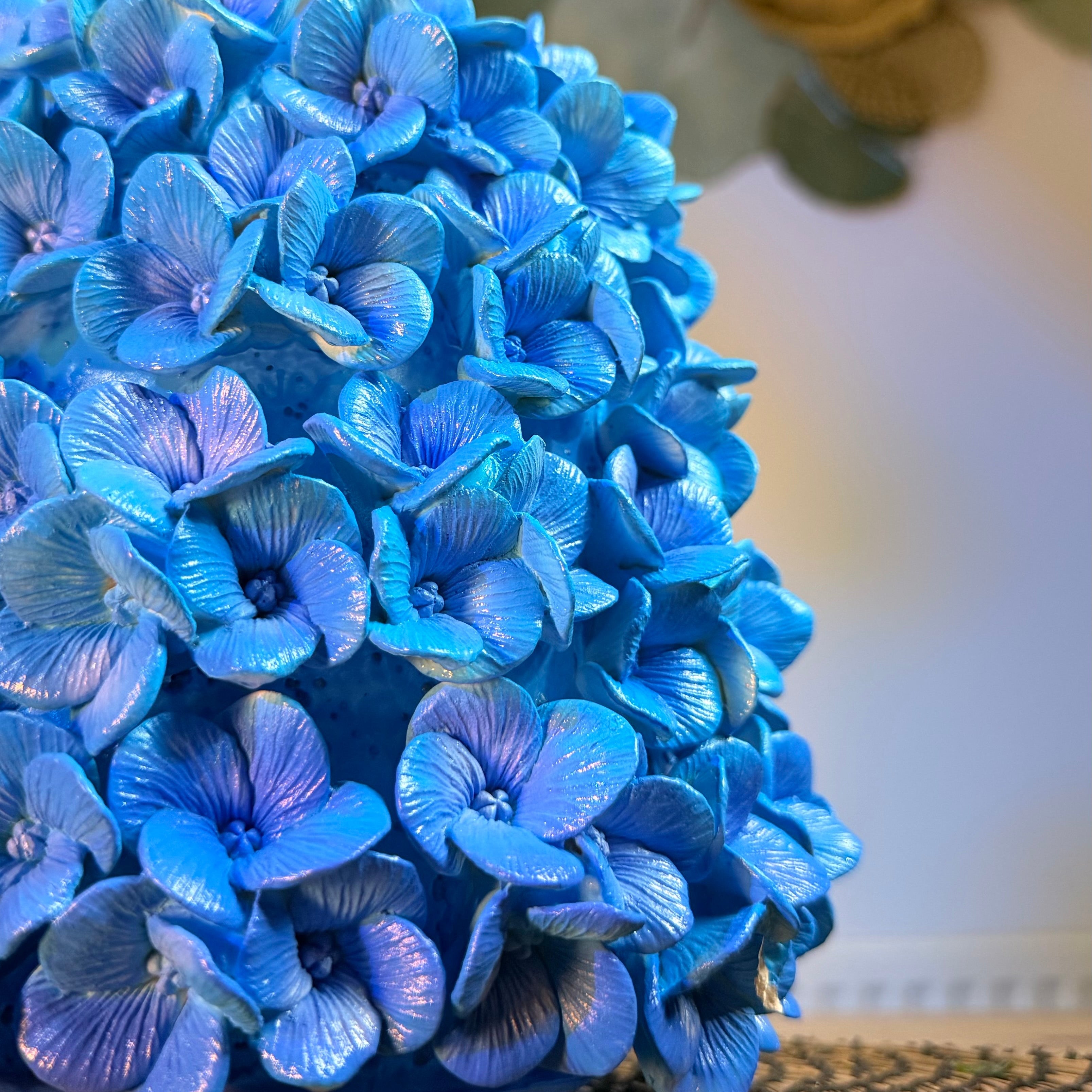 A close-up of a vibrant blue hydrangea flower arrangement on a textured woven surface, with a soft-focus background of green leaves and a warm golden light.