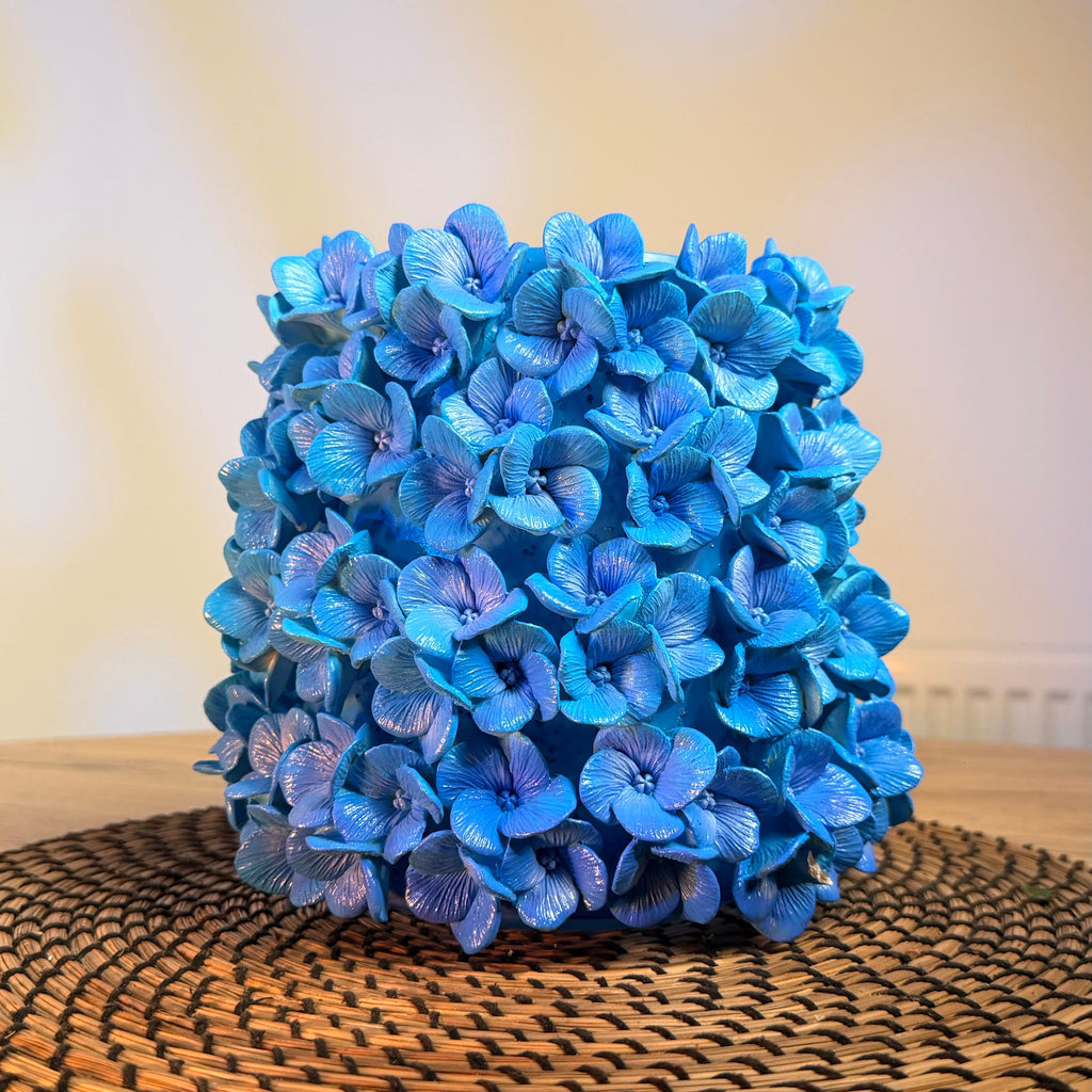 A decorative cylindrical arrangement of bright blue hydrangea-style flowers sits on a round woven placemat over a wooden table, with a softly blurred indoor background.