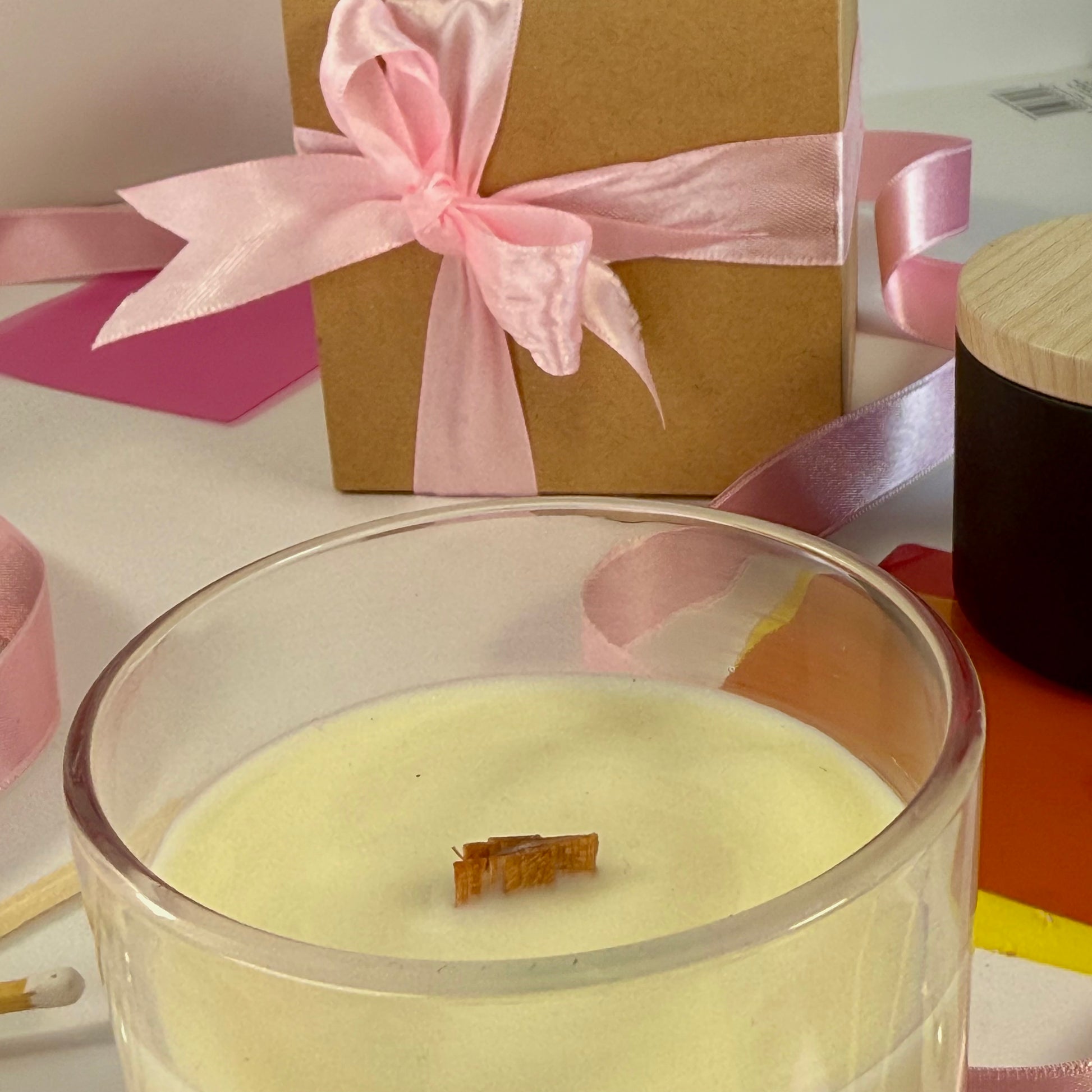 Glass candle with wick on a white surface, with a gift box and pink ribbon in the background.