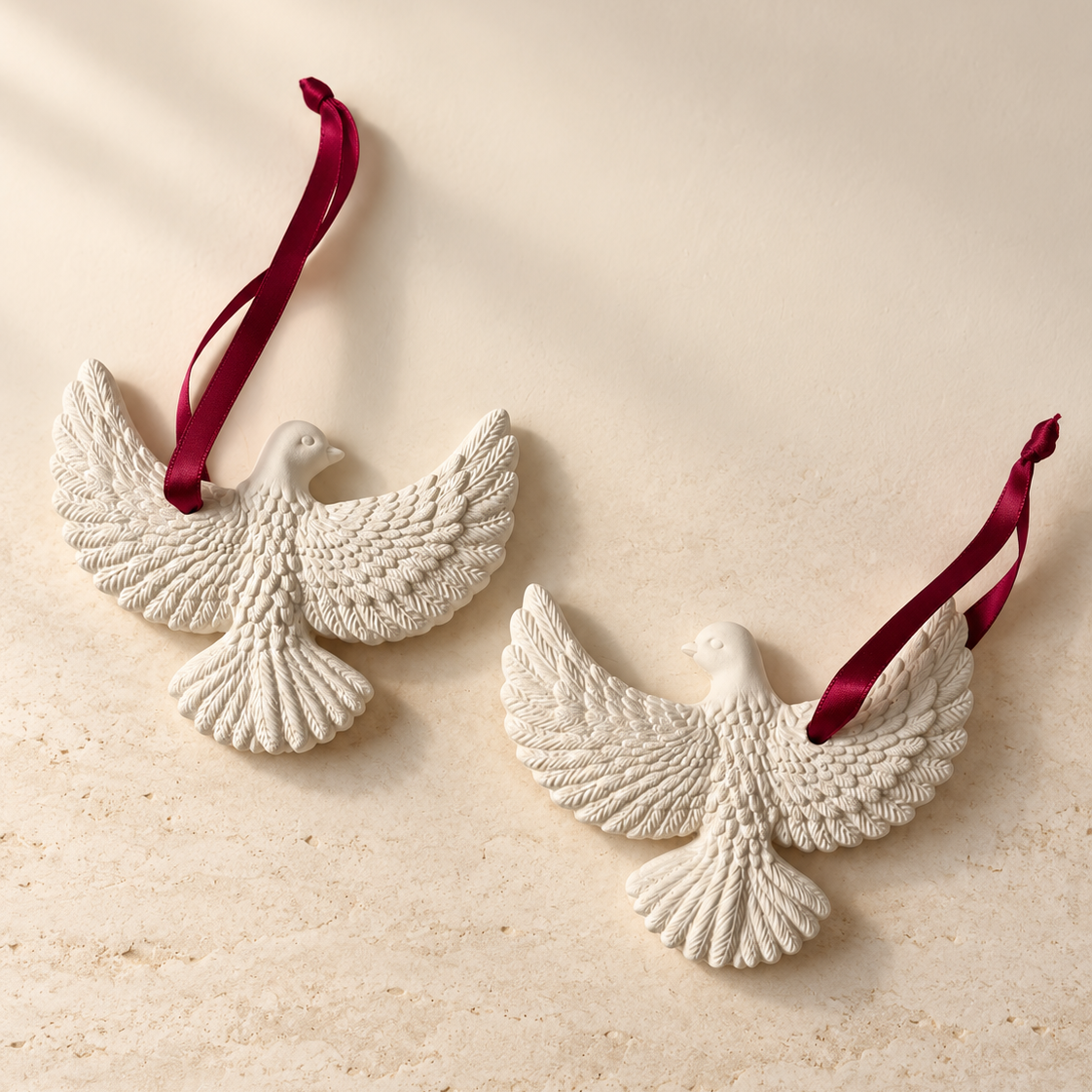 Two white textured dove-shaped ornaments with dark red hanging ribbons, displayed on a warm beige stone-like surface.