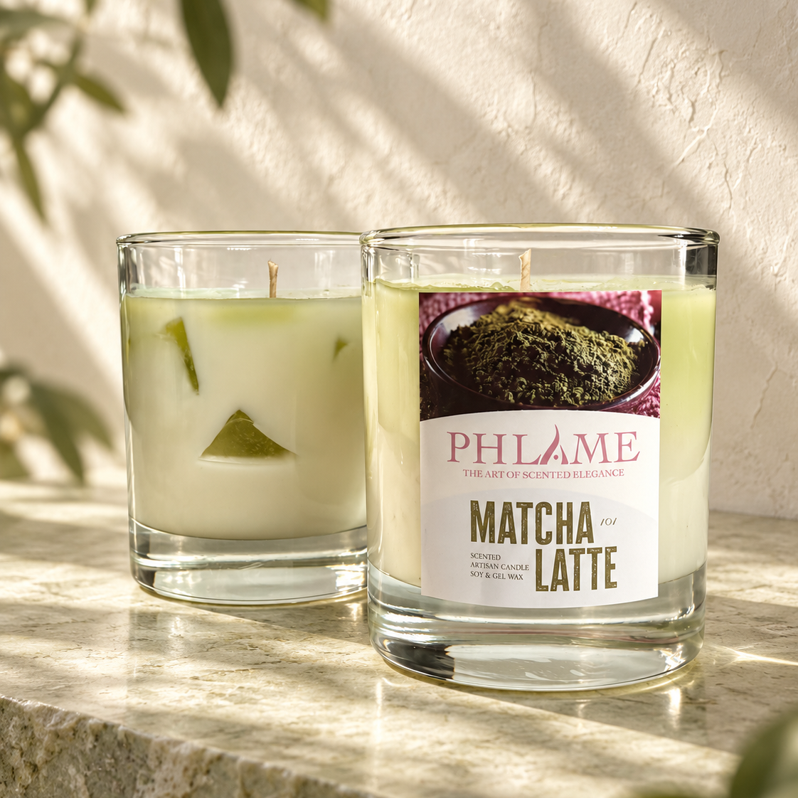 Two PHLAME candles in clear glass jars on a stone surface against a textured wall with leaf shadows. The left candle is pale yellow-green with a green leaf slice inside, and the right candle has a white label reading 'MATCHA LATTE' with a matcha photo and says '100% ORGANIC'.