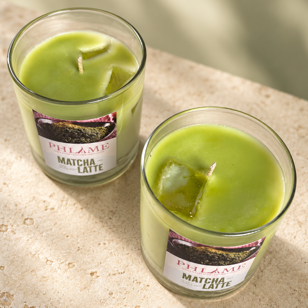 Two green matcha latte scented candles in clear glass jars with white labels, resting on a light beige stone surface with soft natural shadows.