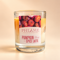 A PHLAME scented candle in a clear glass jar, featuring a label with illustrations of pumpkins and the text 'Pumpkin Spice Latte'. The candle wax is visible through the clear glass, and the background is a soft, warm beige gradient.