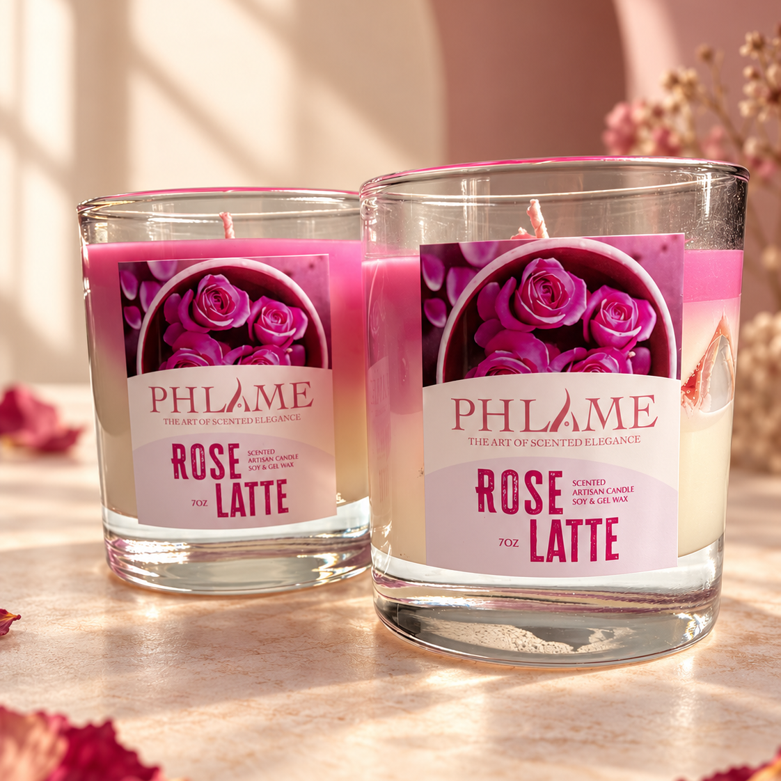 Two PHLAME candles in clear glass jars with pink wax and rose-themed labels, styled on a warm beige surface with scattered dried petals and soft background lighting.