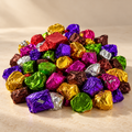 A pile of colorful foil-wrapped wax melts on a light beige surface, wrapped in metallic purple, gold, green, red, brown, silver, and pink foil.