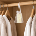 A small clear mesh drawstring bag hanging from a wooden closet rod, containing olive wax sachets. Wooden hangers with neutral-toned shirts are visible on both sides in a softly lit closet setting.
