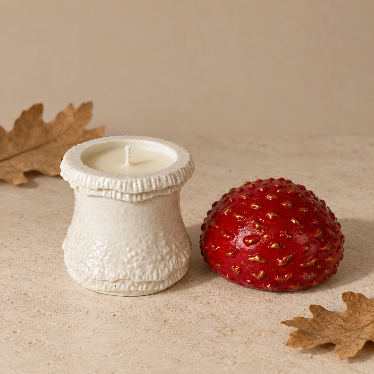 A textured off-white ceramic candle holder with a white wax candle next to a glossy red strawberry-shaped candle, styled with dried autumn leaves on a beige stone surface against a neutral beige background.