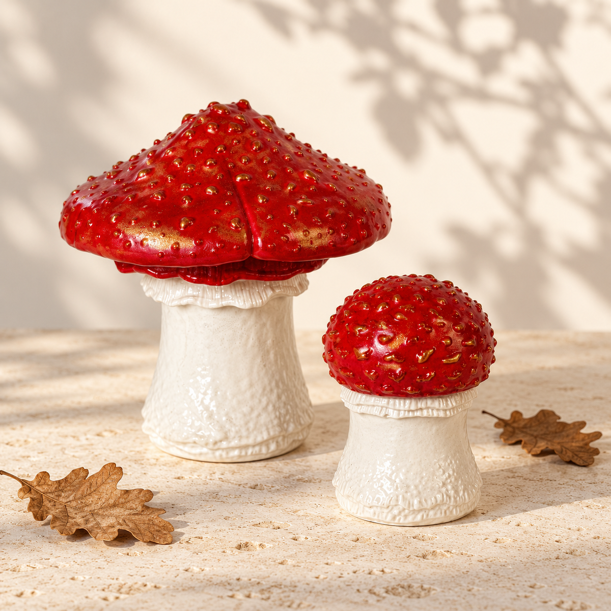 Two red-and-white mushroom-shaped decorative objects on a light stone surface with dried oak leaves and soft tree shadows in the background.