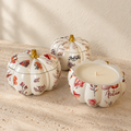 Three decorative pumpkin-shaped candle holders with a white base and red and orange floral print, arranged on a textured beige stone surface with warm sunlight and shadows.