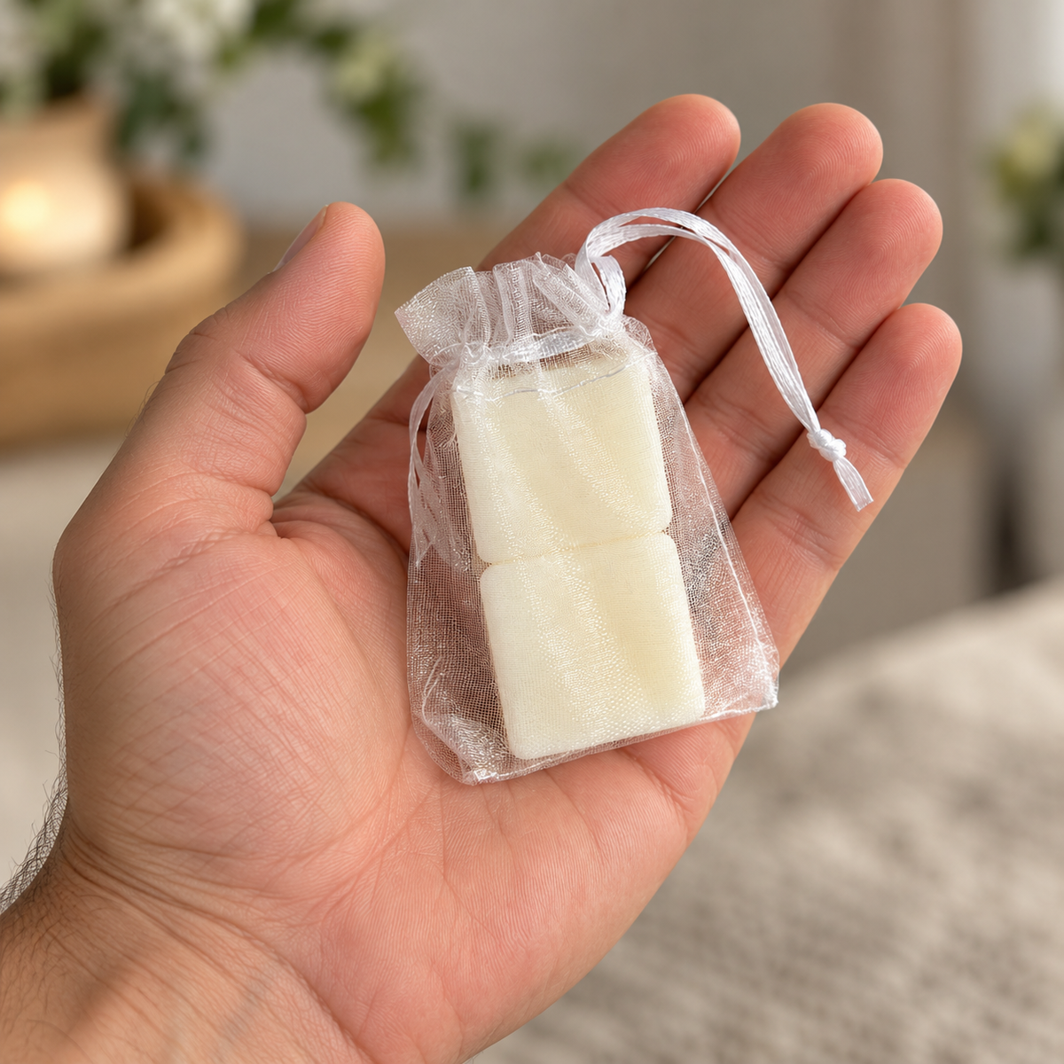 A hand holding a small clear organza drawstring pouch containing two olive wax sachets, with a softly blurred indoor background.